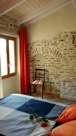 Une chambre avec un mur en pierre, une chaise en bois, un lit recouvert d’une courtepointe et une fenêtre avec des rideaux.