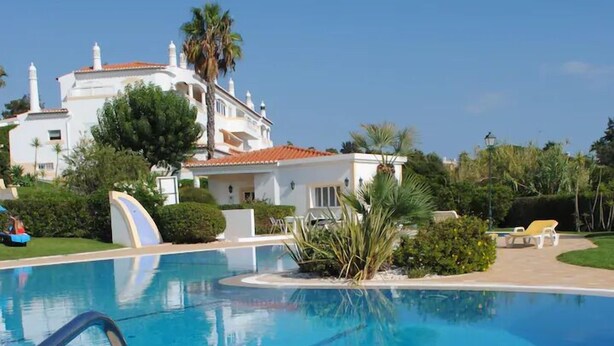 A swimming pool with a clear blue water, surrounded by a tiled deck and a white building with a red roof in the background. - Beautiful Residential Villa on An Exclusive Private Complex, pool, roof terrace (Carvoeiro)