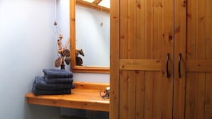 A wooden wardrobe with two handles, a mirror above a wooden shelf with folded towels, and a small stool.