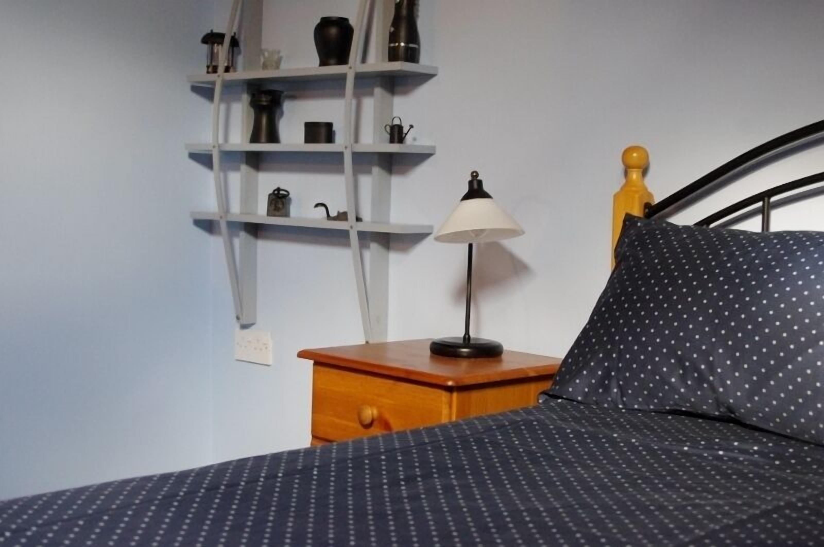 A bed with a patterned bedspread, a bedside lamp, a wooden nightstand, and a shelf with various items.