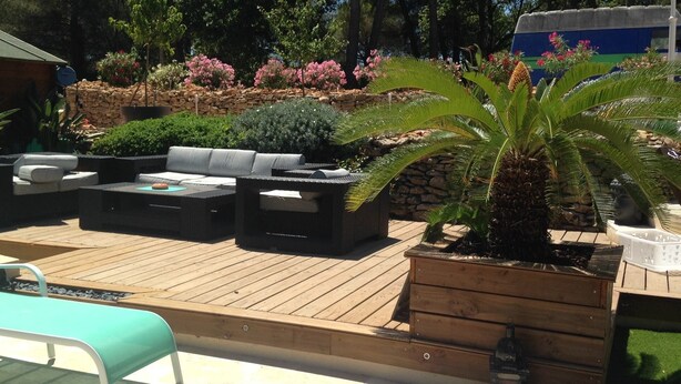 Een terras buiten met een houten terras, een hoekbank en een palmboom.