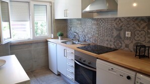 A compact kitchen with a sink, stove, and dishwasher, featuring a patterned backsplash and a window with blinds. - Lisbon Beach Apartments 3. Beach 5 minutes, terrace, barbecue, pets welcome (Costa da Caparica)
