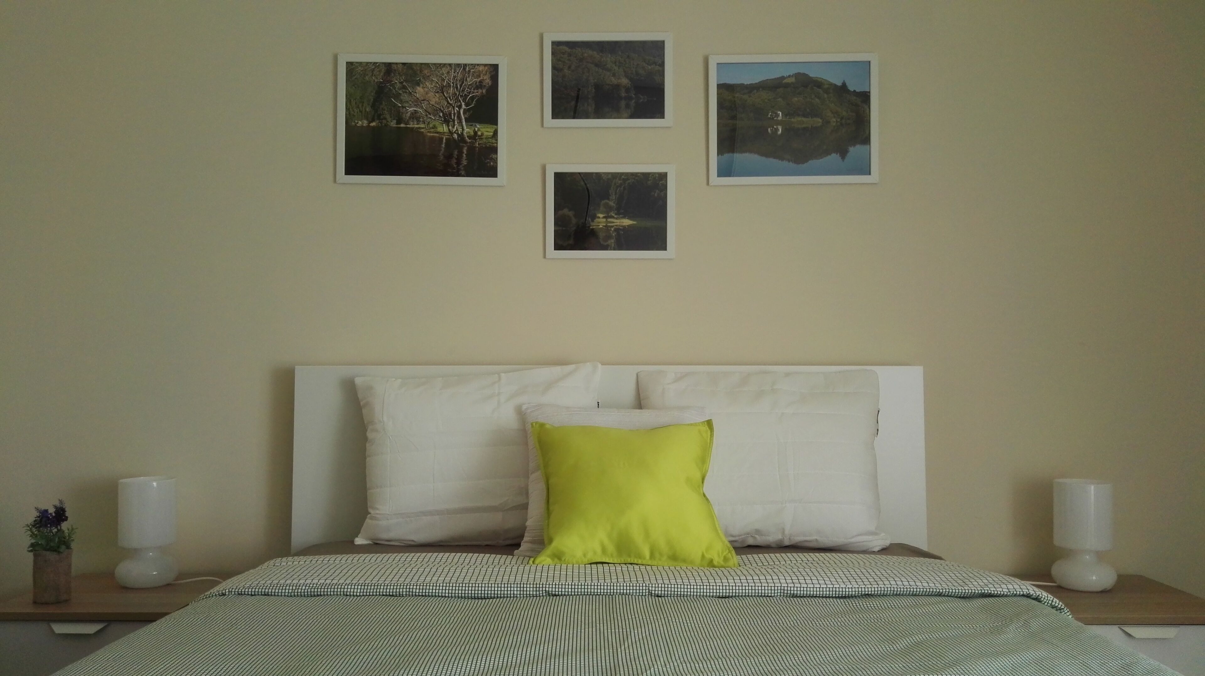 A neatly made bed with a patterned bedspread, a white headboard, and a single bright green pillow. There are two bedside tables with white lamps and a small potted plant on one. Four framed pictures are hung on the wall above the bed.