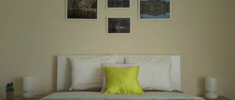 A neatly made bed with a patterned bedspread, a white headboard, and a single bright green pillow. There are two bedside tables with white lamps and a small potted plant on one. Four framed pictures are hung on the wall above the bed.