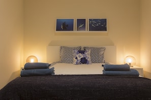 A neatly made bed with a dark cover, two pillows with a floral pattern, and two bedside lamps. Above the bed, there are three framed pictures depicting underwater scenes.