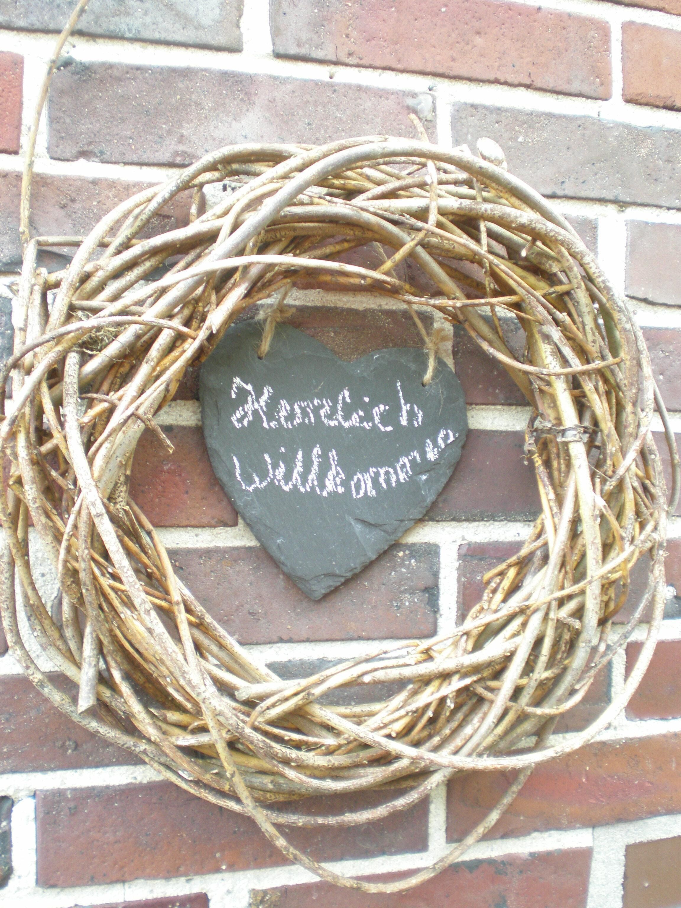Een hartvormig schoolbord met de tekst 'Herzlich Willkommen' tegen een bakstenen muur, omringd door een geweven krans.