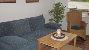 Un salon avec un canapé bleu, une table en bois sur laquelle est posée une bougie, un fauteuil en osier et une fenêtre avec des stores.