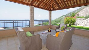 Un espacio al aire libre techado con muebles de mimbre, una mesa con una bebida y vistas al mar y las colinas.
