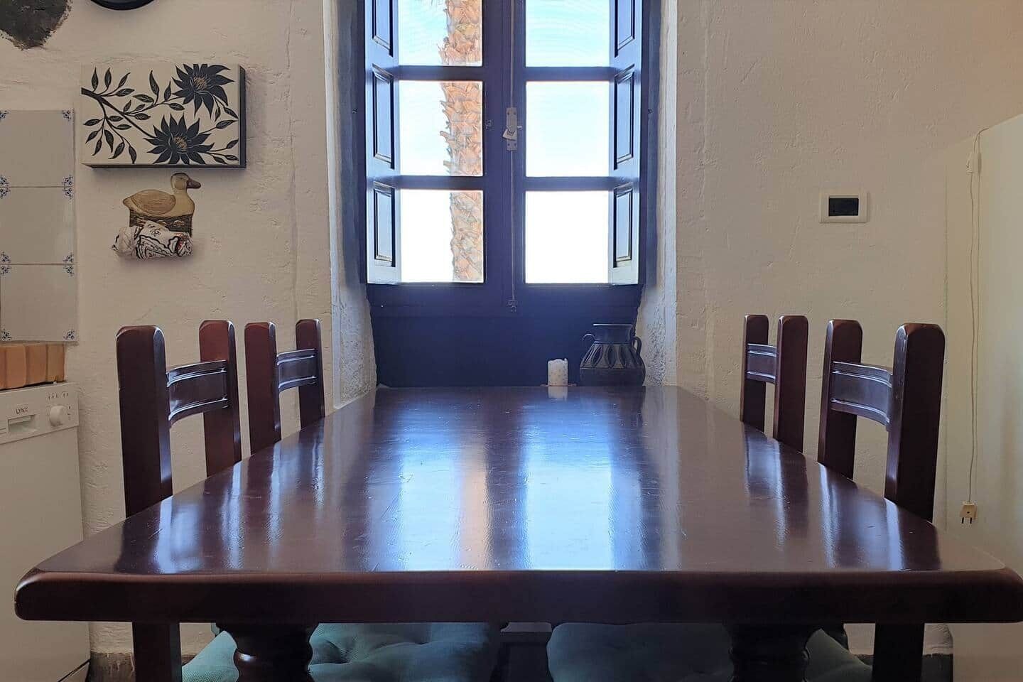 Een eetkamer met een grote houten tafel en zes stoelen. Er is een raam met veel ruitjes en uitzicht op het omliggende landschap.