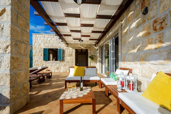 A covered outdoor seating area with stone walls, a wooden table with a lace doily, and a bench with cushions. - Villa Panoramic View by MyWaycation (Dubravka)