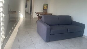 A living room with a grey sofa, a wooden table, and a chair. There is a kitchen in the background with a dining table and chairs.