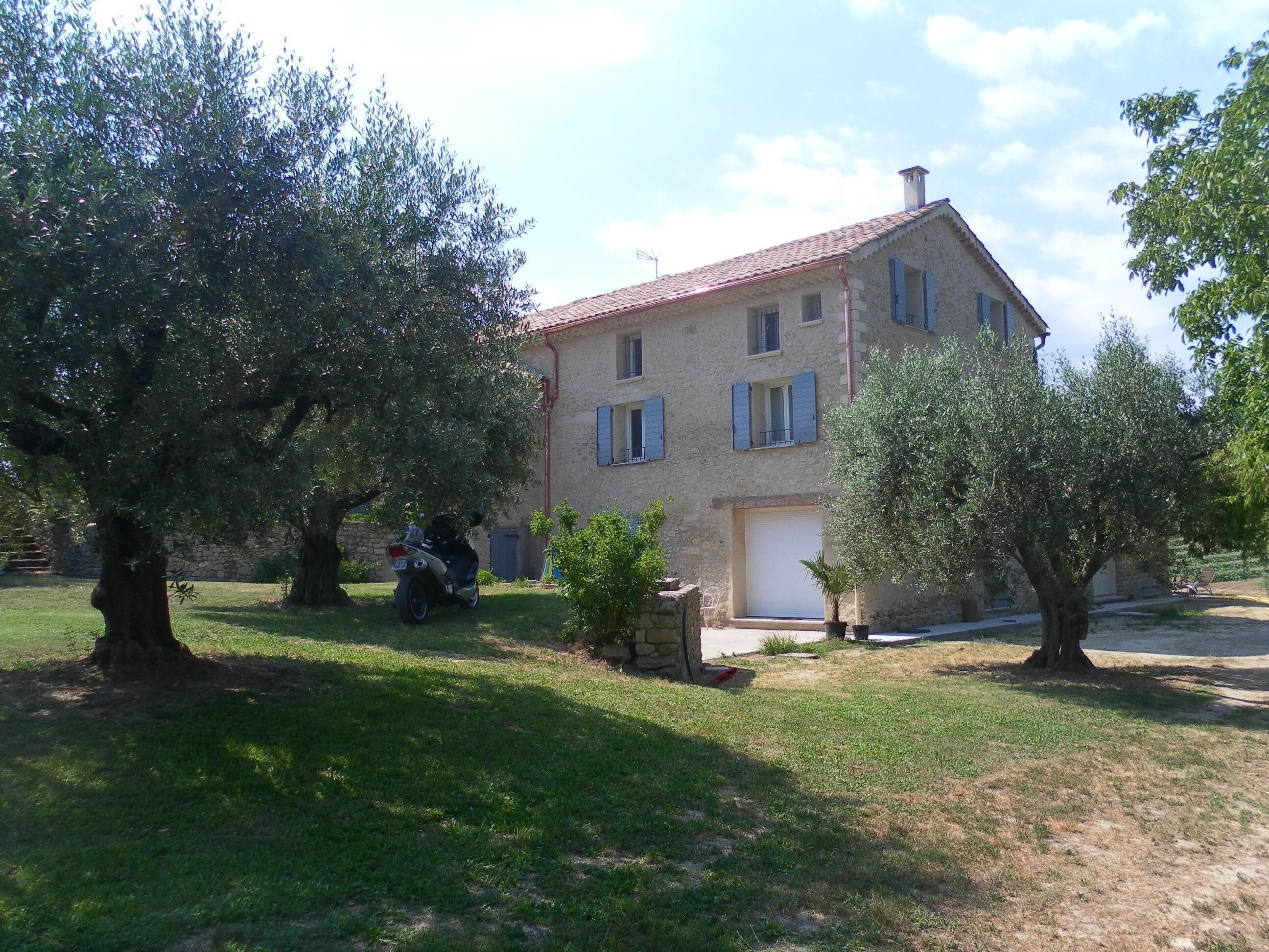 A two-story stone building with a tiled roof, a garage, and a well-maintained lawn with olive trees.