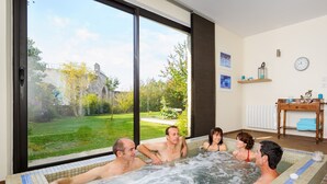 A hot tub with four people, a large window, and a view of a garden and building.