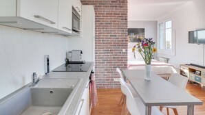 Een moderne keuken met een accentmuur van baksteen, een eetgedeelte met een tafel en stoelen, en een slaapkamer met een bed en een televisie.