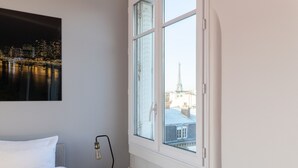 Hotelkamer met een bed, nachtkastje met lamp, radiator en raam met uitzicht op stadsgezicht en een toren.