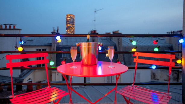 Een rode metalen tafel met twee champagneglazen en een metalen emmer, op een balkon met rode metalen stoelen en kleurrijke lichtslingers.