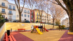 A playground with a slide and climbing structures, surrounded by a red safety surface, benches, and trees.