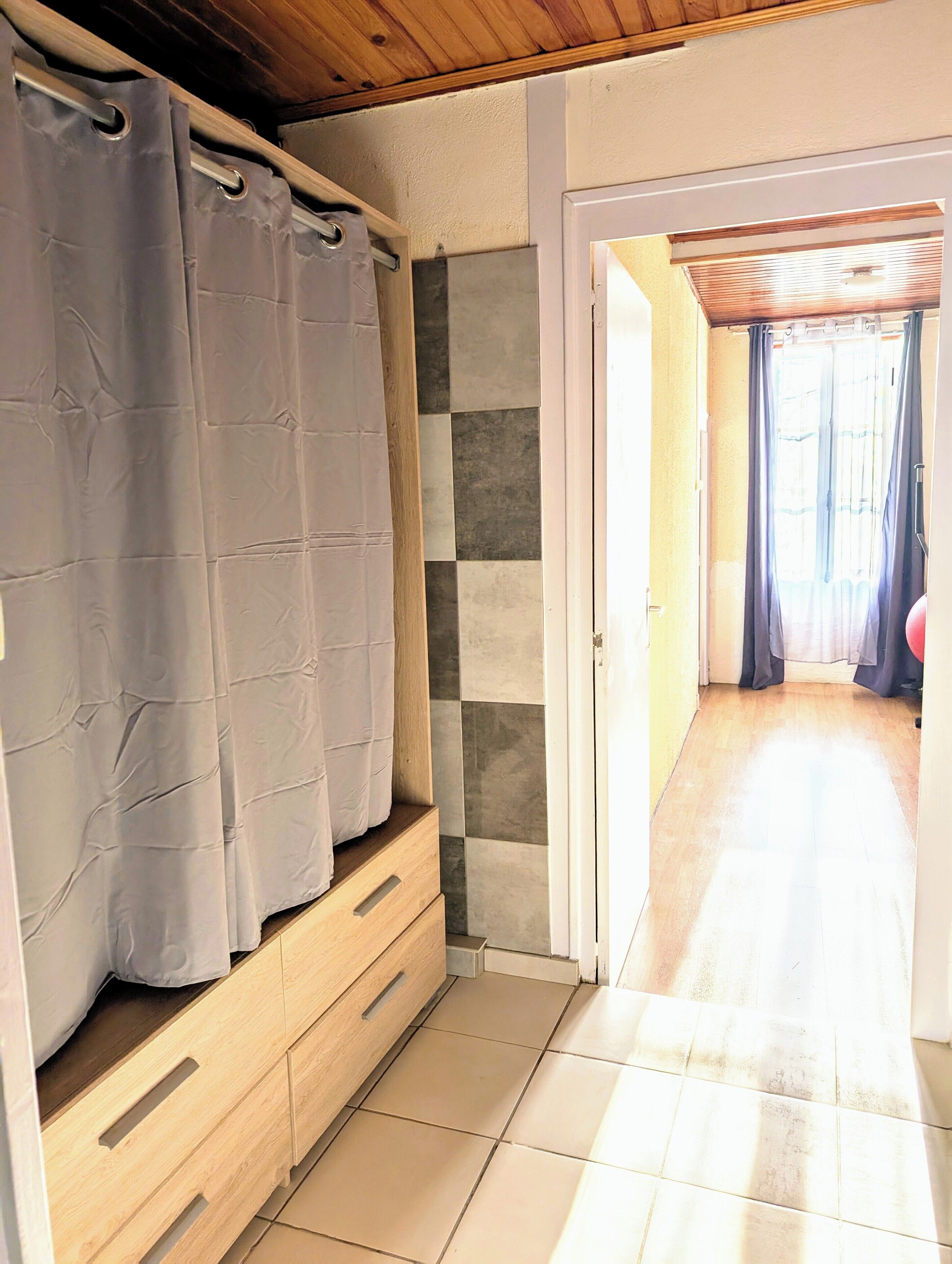 Una habitación con techo de madera, zona de ducha con cortina, suelo de baldosas y vista a un pasillo con puerta al exterior.