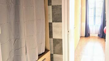 Una habitación con techo de madera, zona de ducha con cortina, suelo de baldosas y vista a un pasillo con puerta al exterior.