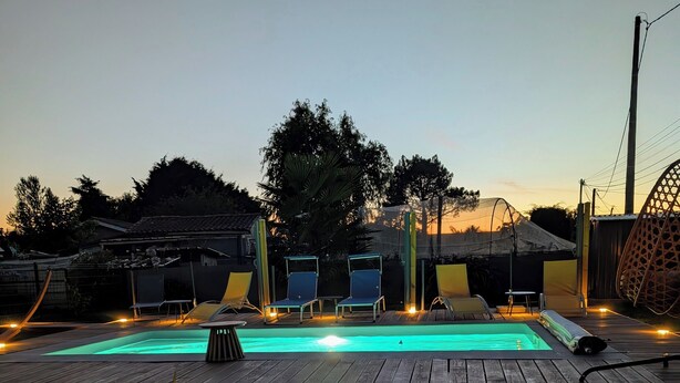 A wooden deck leading to a swimming pool with a clear blue water feature, surrounded by a fence and equipped with outdoor seating and lighting. - L'échappée Huroise, private pool and spa (Hure)