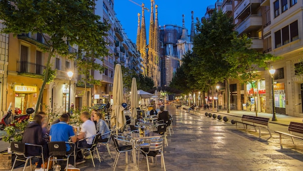 Ein Straßencafé mit Tischen und Stühlen, umgeben von Bäumen und Gebäuden.