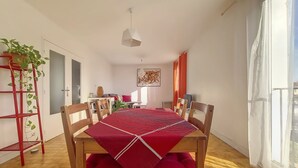 Un coin repas avec une table et des chaises en bois, une nappe rouge et une plante en pot posée sur une étagère en métal rouge.