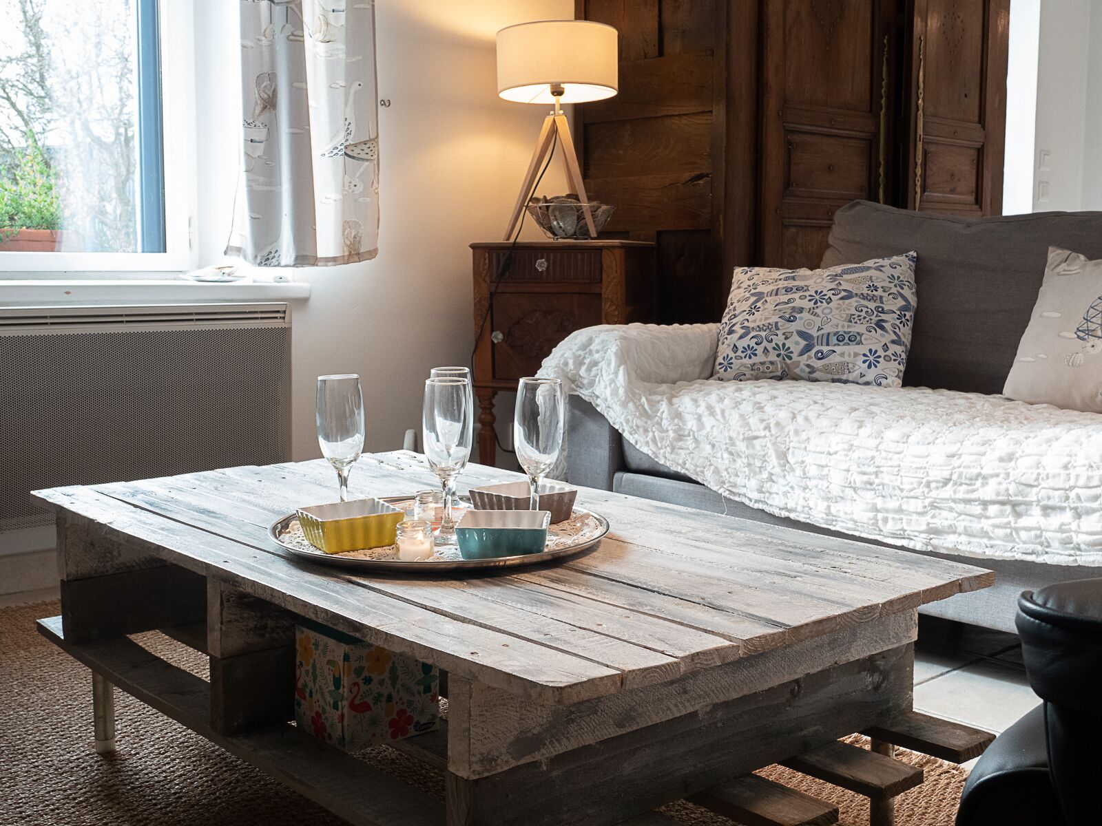 A rustic wooden table with a tray of glasses and bowls, a lamp, and a bed with patterned pillows in a cozy room with a window and wooden walls.