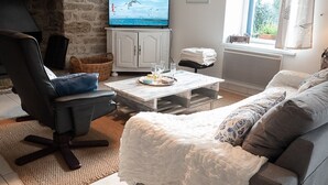 A cozy living room with a stone wall, a television, a white cabinet, and a window with curtains. - Fisherman's cottage in St Guénolé, near Pors Carn, La Torche (Penmarch)