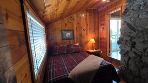 Una habitación de cabaña acogedora, con paredes de madera, una colcha escocesa, una mesita de noche con una lámpara y una pared de piedra.
