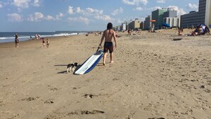 A sandy beach with footprints, a person carrying a surfboard, and buildings in the background.