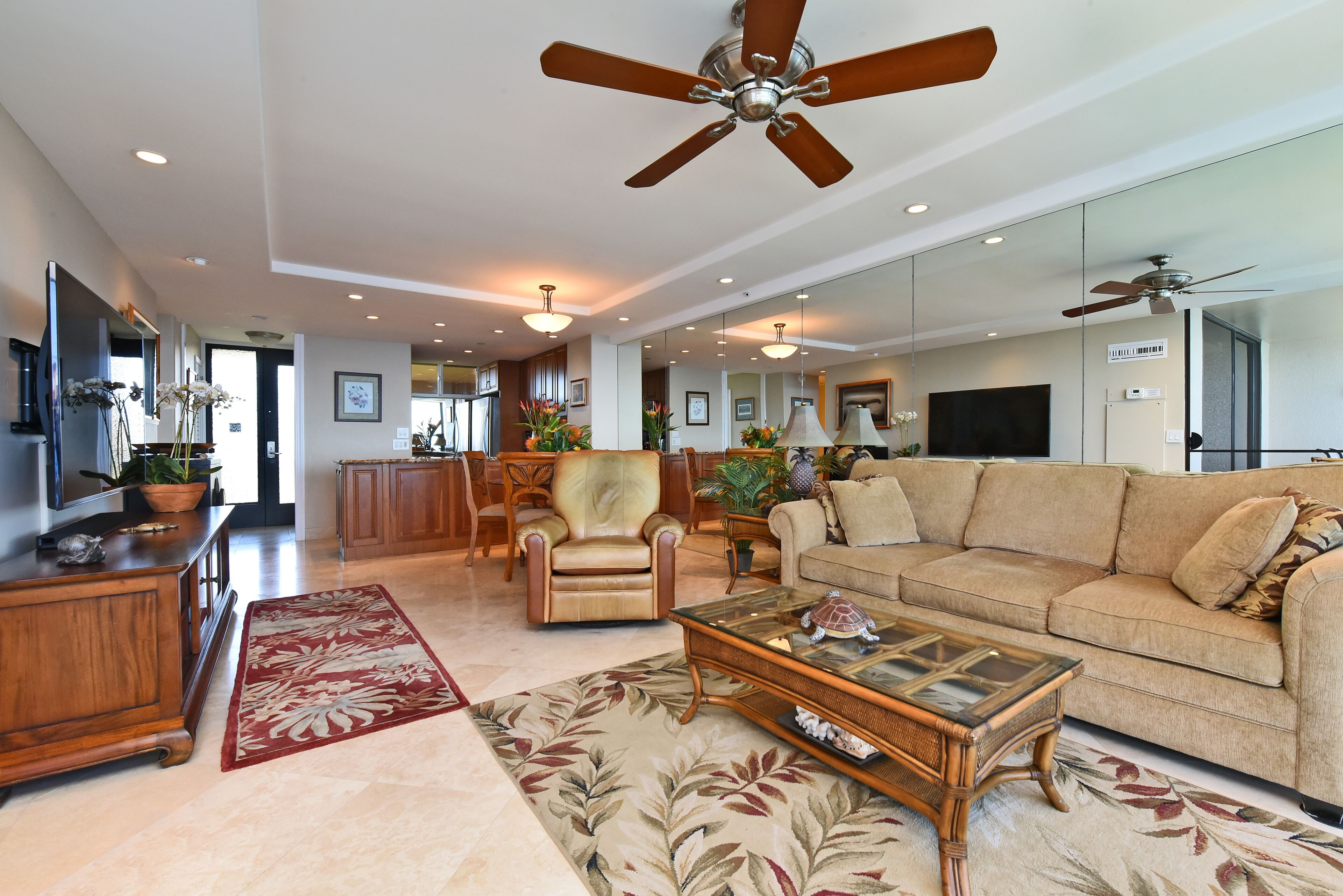 A spacious living room with a large sofa, a coffee table, and a fireplace. There are ceiling fans, a TV, and a dining area visible.