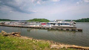 A docked houseboat with a covered deck area, a small boat, and a pier extending into a lake with a forested shoreline. - Bourbon Trails, Fishing and Relaxing - Taylorsville Lake has it all! (Taylorsville)