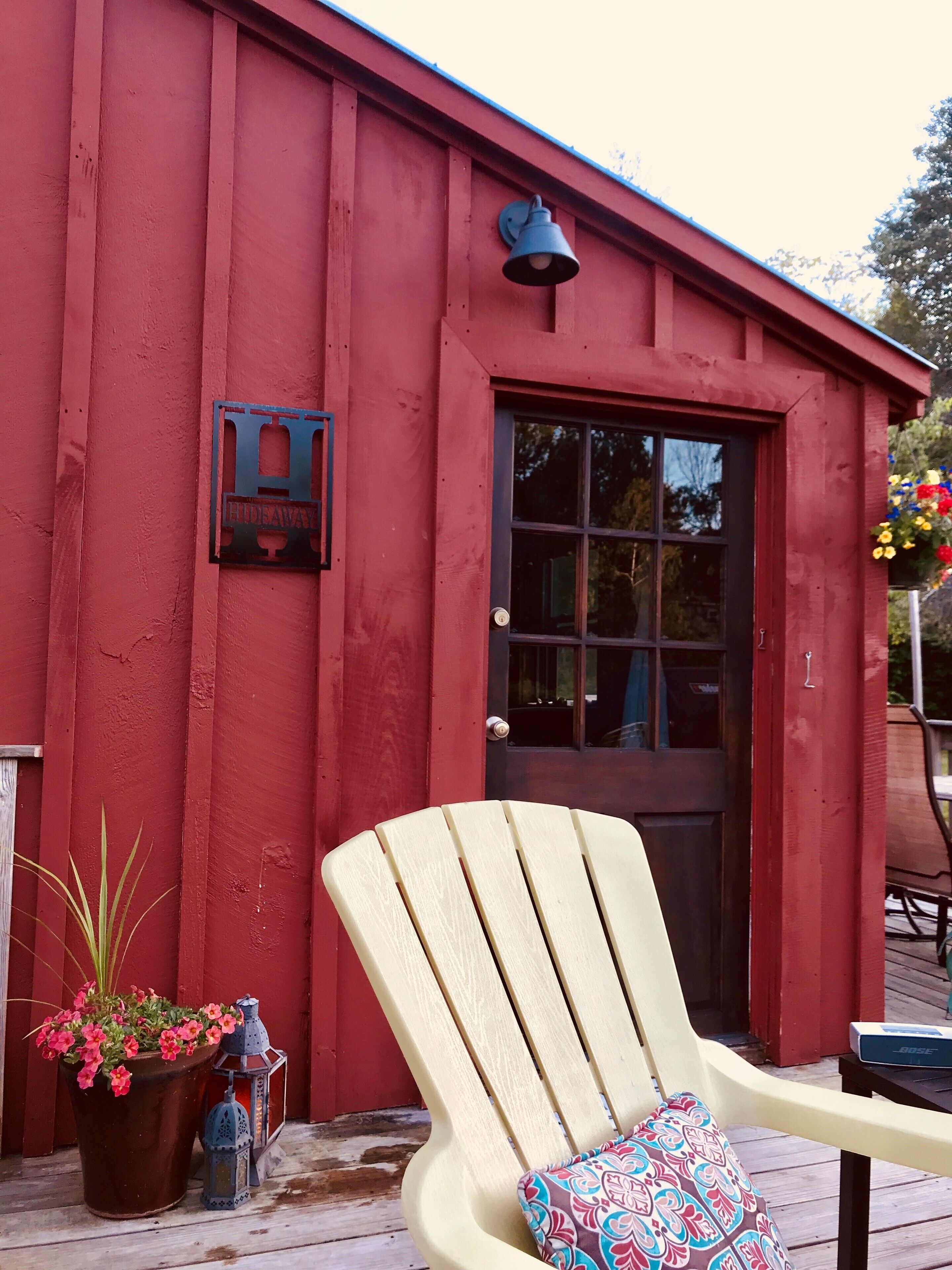 A red building with a black door, a chair with a patterned cushion, a potted plant with pink flowers, and a lantern.