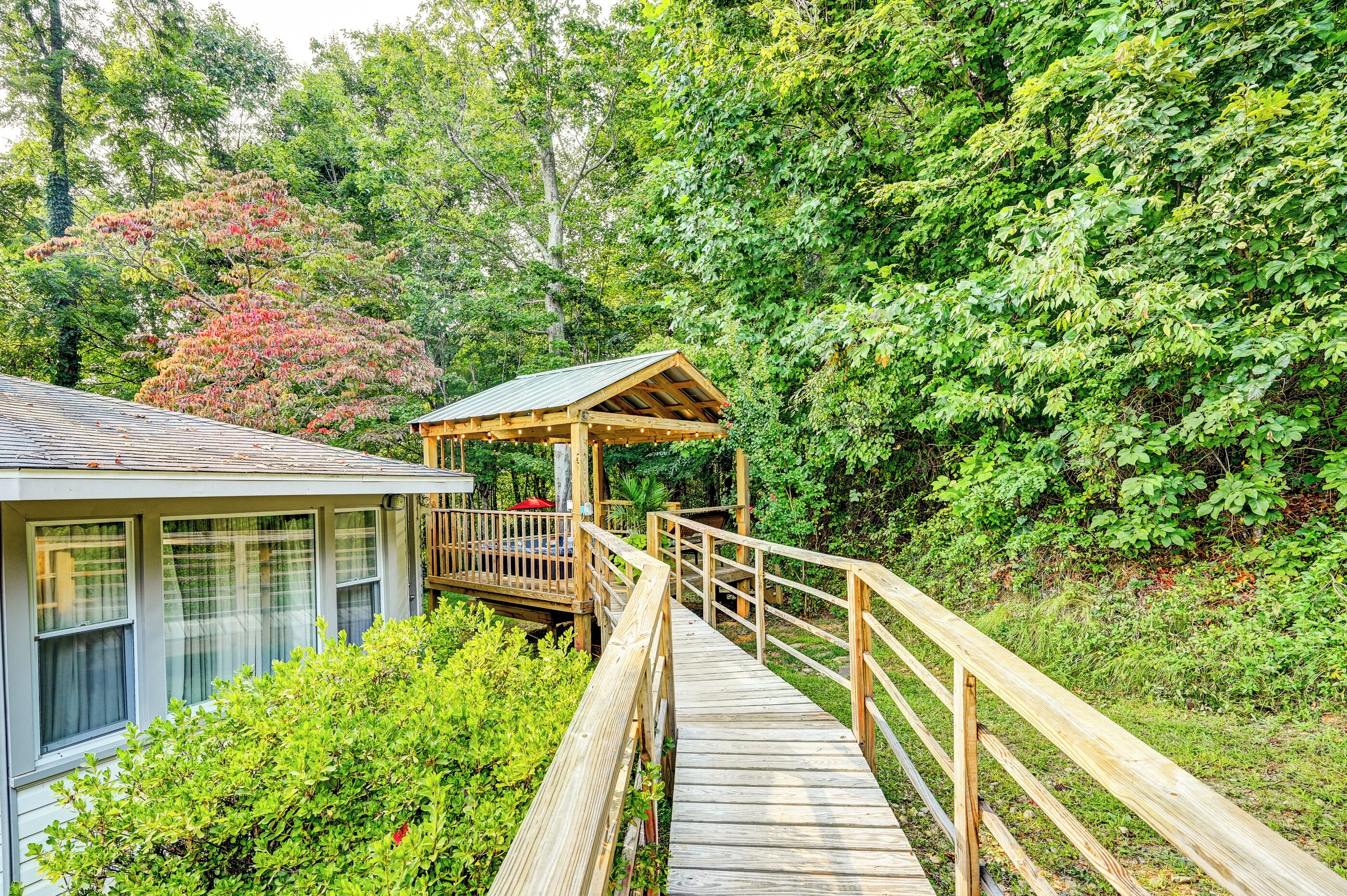 A wooden deck leading to a covered gazebo with a metal roof, surrounded by lush greenery.