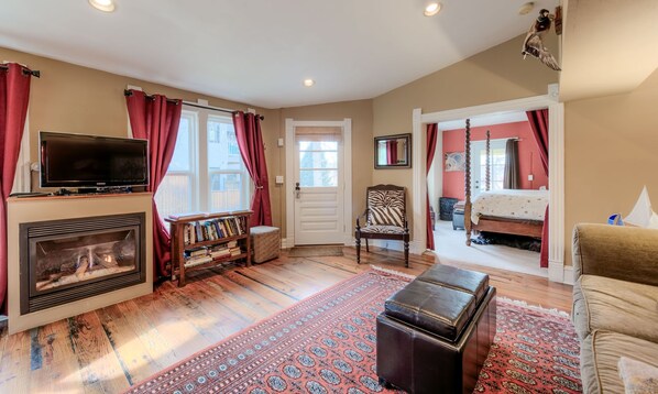 A cozy living room with a fireplace, a television, a bookshelf, and a large window with curtains.