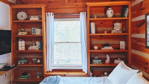 A wooden cabin bedroom with a bed, built-in bookshelves, and a window with curtains.