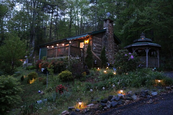A rustic cabin with a stone chimney, surrounded by a garden with various plants and flowers, and a gazebo in the background.