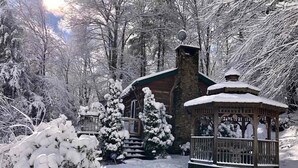 A snow-covered lodge with a stone chimney, a wooden gazebo, and snow-laden trees.