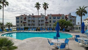 Uma área de piscina com espreguiçadeiras e guarda-sóis, um edifício com varandas e palmeiras.