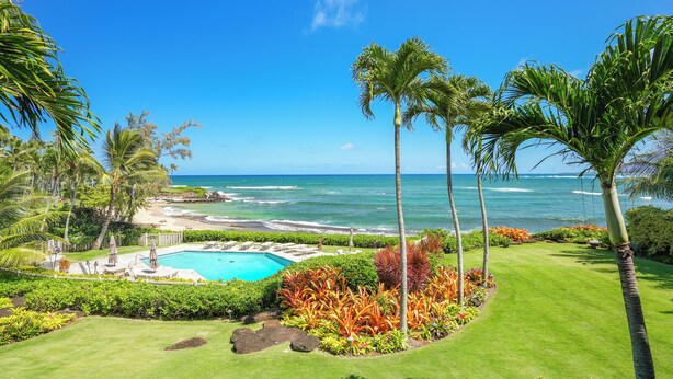 A pool with a view of the ocean, surrounded by palm trees and a well-maintained lawn. - Lanikai 232 “Seascape” | Ocean View | Central A/C | Sleeps 4 (Kapaa)