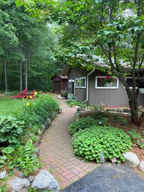 A garden pathway leading to a house with a garage, surrounded by lush greenery and flowering plants. - Thunderbird Lodge: Sandy Beach, Pontoon Boats & Northwoods Memories (Lac du Flambeau)