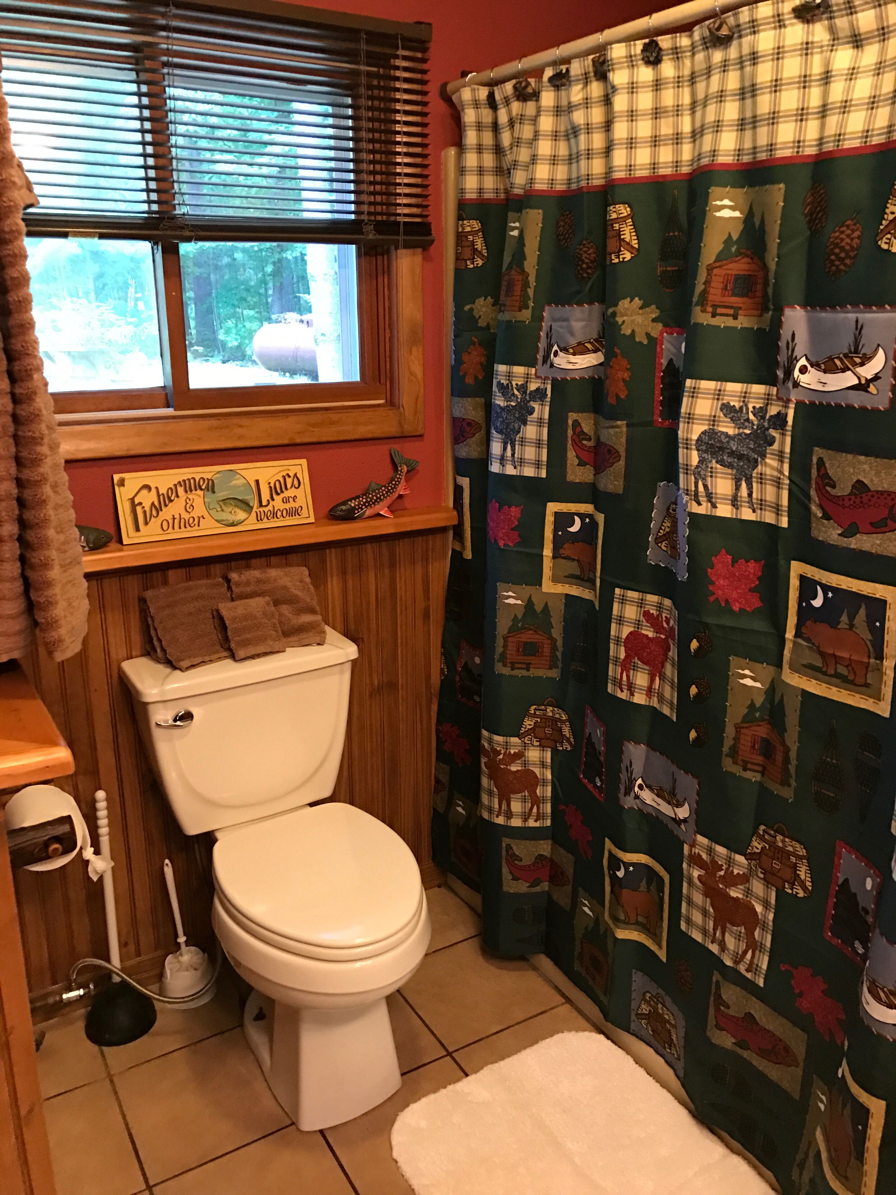 Un baño con un inodoro, una ventana con persianas, una cortina de ducha con estampado de animales y un bosque, y un cartel en la pared.