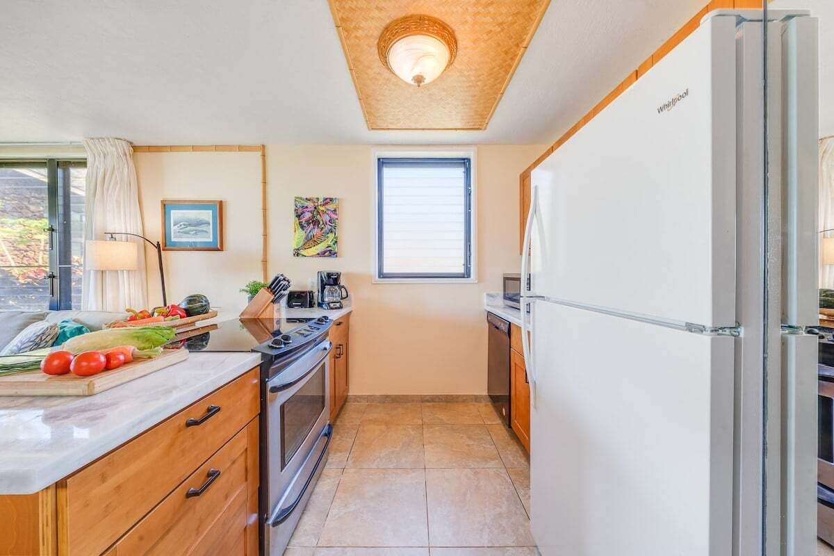 A compact kitchen with a stove, oven, microwave, and refrigerator. There is a window with blinds, a wooden ceiling, and a countertop with vegetables.