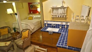 A hotel room with a bed, a sitting area with chairs, a small table, a kitchen area with a sink and a tiled backsplash, and a shelf with dishes and glasses. - Cozy Studio Apartment with European style (Laguna Beach)