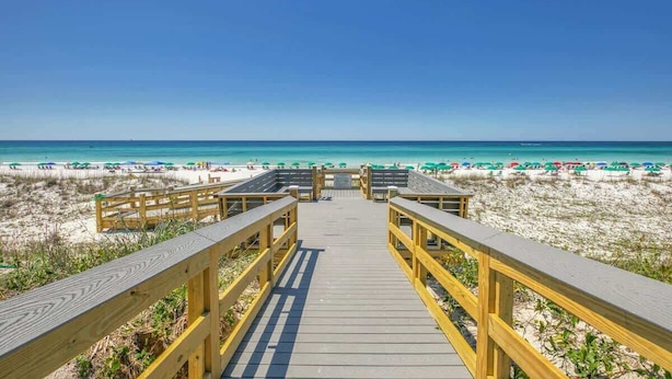 A wooden boardwalk leading to a beach with umbrellas and beachgoers.