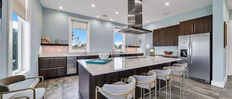 A modern kitchen with a central island, bar stools, stainless steel appliances, and a view of the outdoors.