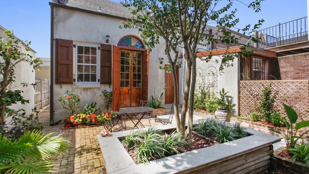 Une maison avec une porte en bois, un petit patio avec une table et des chaises, et un jardin avec différentes plantes.
