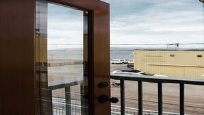 Une vue depuis un balcon, avec une porte en bois, une rambarde en métal, et on aperçoit un parking avec des voitures et un bâtiment en arrière-plan.