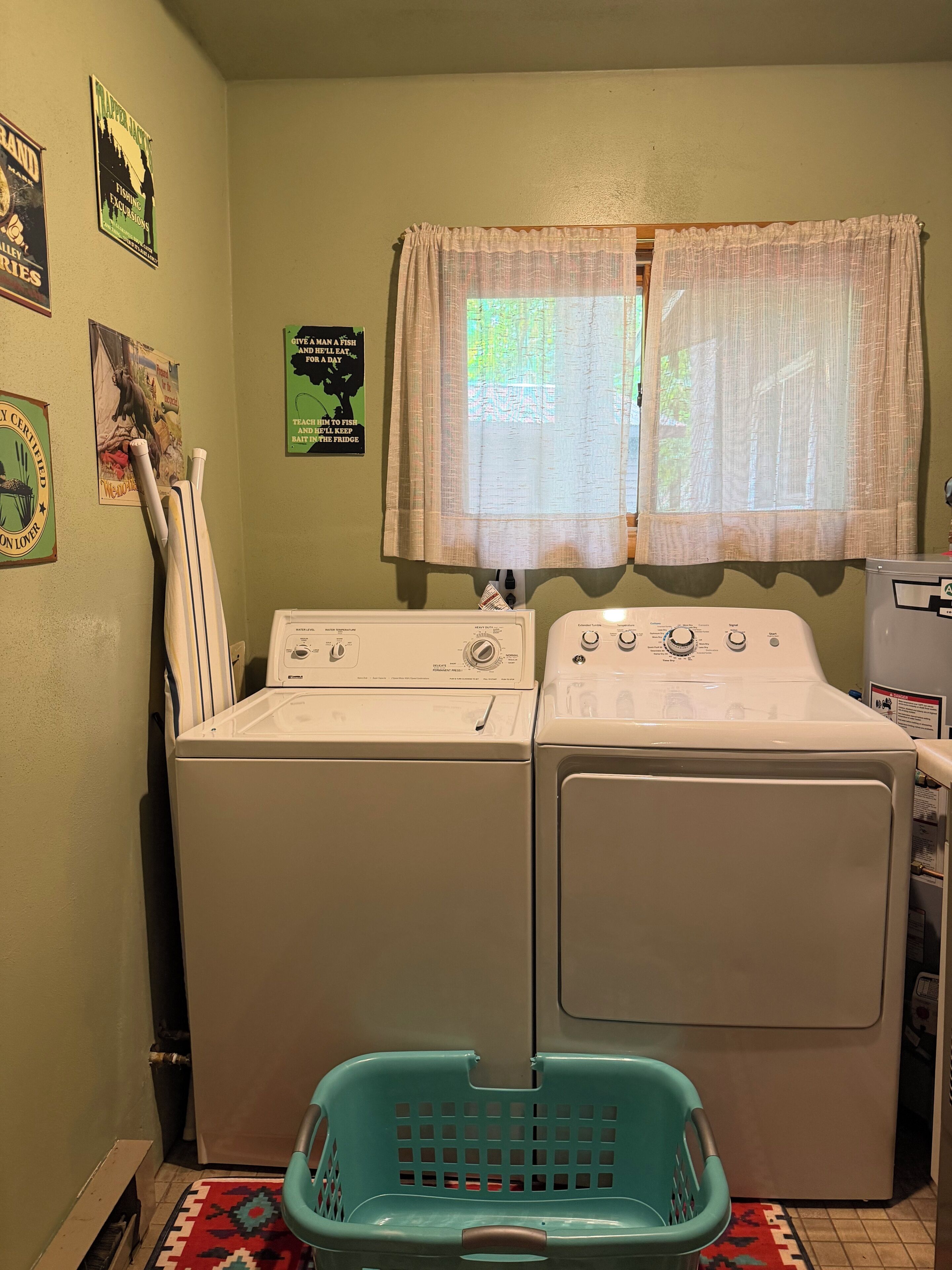 Un cuarto de lavado con dos lavadoras y secadoras blancas, una cesta de ropa azul y una ventana con cortinas traslúcidas.