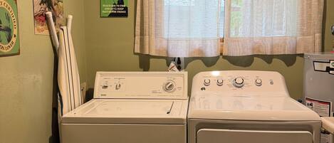 Un cuarto de lavado con dos lavadoras y secadoras blancas, una cesta de ropa azul y una ventana con cortinas traslúcidas.
